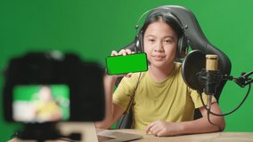 Asian Kid Girl Holding Mobile Phone With Mock Up Green Screen And Talking To Camera While Live Stream In Green Screen Studio
