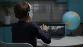School boy playing video game on laptop using joystick sitting at home. Kid in headphones playing online racing game using laptop.