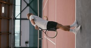 Vertical video of a tennis workout on an indoor tennis court. A young man of European appearance throws a tennis ball with a racket over the net