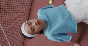 Young woman tennis player of Asian appearance sits on the floor in the middle of a tennis court and looks directly at the camera without smiling, close-up, vertical video