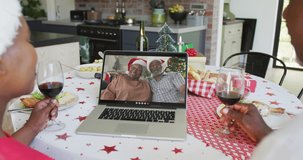 African american couple with wine using laptop for christmas video call with couple on screen. christmas, festivity and communication technology.