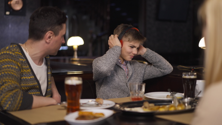 Misbehaving teenage son putting on headphones as parents scolding boy sitting in restaurant. Unhappy Caucasian teenager with father and mother dining in cafe indoors. Slow motion