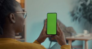 Young woman using smartphone with green empty screen for video call at home. African american female in eyeglsses sitting on couch with modern gadget in hands.
