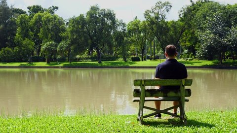 Mental health concept of loneliness, depression, sad emotional, mental illness. Back view of sad thoughtful unhappy man sitting alone on bench near lake in green city park. Heartbreak, broken love