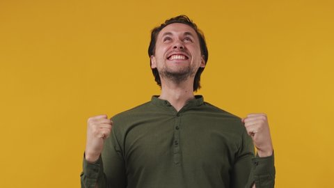 Excited jubilant overjoyed happy joyful happy smiling young brunet bearded man 20s wears green shirt doing winner gesture celebrate clenching fists say yes isolated on pastel plain yellow background