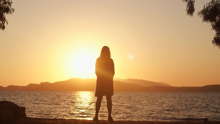 Dark silhouette of adult european woman standing alone in scenic sunset marine landscape background. 4k video of sunny orange sky, mountains, sea water, trees and woman