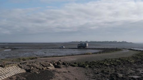 Distant view of mysterious Osea Island private retreat causeway with moving vehicles. Low tide river Blackwater estuary. Overcast winter day outdoors. United Kingdom, Essex, Maldon, November 11, 2021