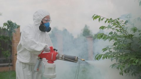 Farmer agronomist in protective suit and respirator treats fruit trees from pests and diseases using fogger machinespraying smoke with organic pesticides in garden.