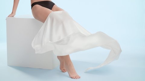 Good-looking slim woman in black underwear throws a piece of white silk cloth down her smooth legs sitting on white cube platform on pale blue background | Laser hair removal commercial