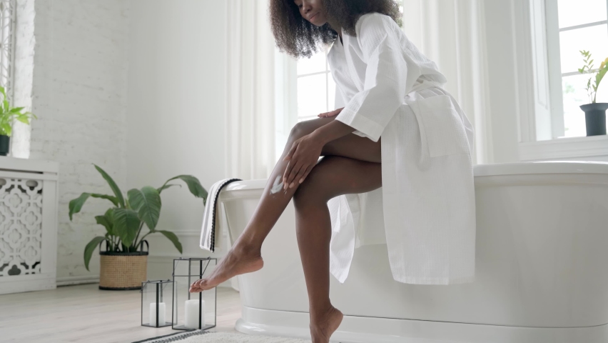 Young gorgeous African American black woman wearing white gown applying putting replenishing lotion on dry legs skin sitting on bath tube in bathroom. Skincare, self love, body care spa concept.