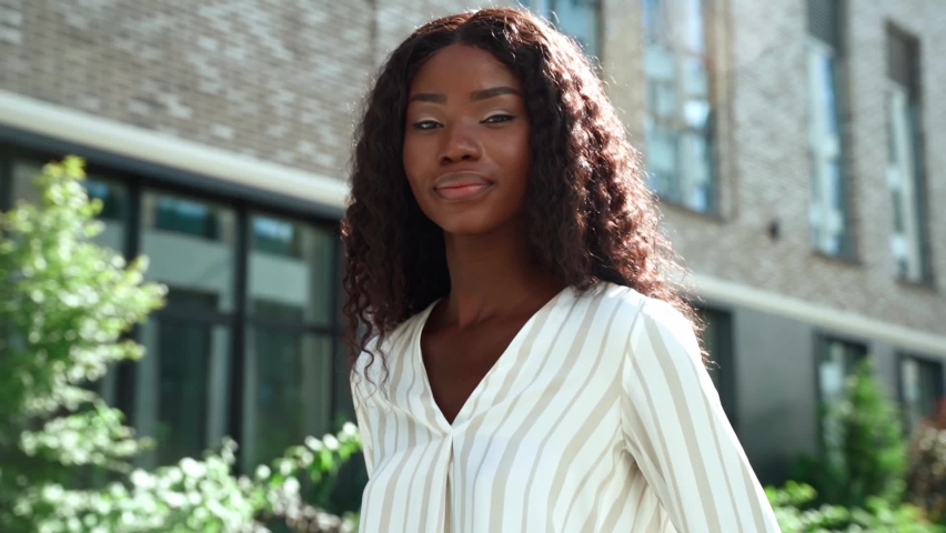 Happy smiling young adult ethnic black African attractive millennial business woman or student with curly long hair posing standing outdoor on sunny city street looking at camera. Slow motion portrait