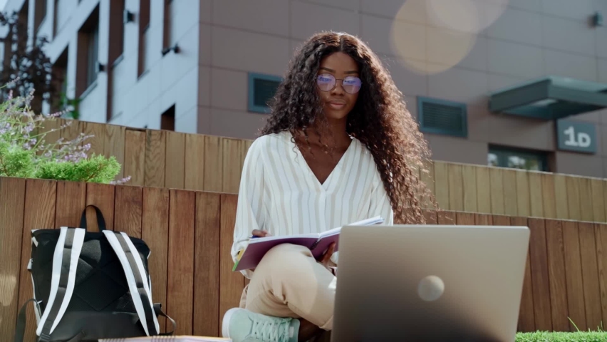 Young black African woman university student learning online using laptop computer, taking notes, watching webinar or virtual education remote class studying outdoor sitting outside uni campus area. Royalty-Free Stock Footage #1082255426