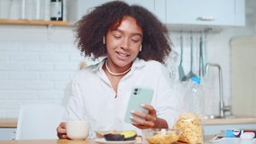 Confident young African American woman talking by video call during breakfast. Successful Hispanic female in kitchen with phone in hand. Businesswoman home video conferencing, business, technology