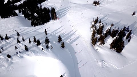 Aerial view of ski resort with people snowboarding down the hill. Stock footage. Flying over the ski or snowboard track on white snow surrounded by dense forest in winter season, travel and sport