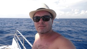 Man in summer hat taking selfie video on catamaran yacht with caribbean sea water background. Summer holidays. Travel vacation
