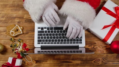 Top view Santa hands in white gloves typing on the keyboard by wooden New Year decorated table. Santa Claus works with laptop, looks through mail and prepares gifts for children. Close up. Slow motion