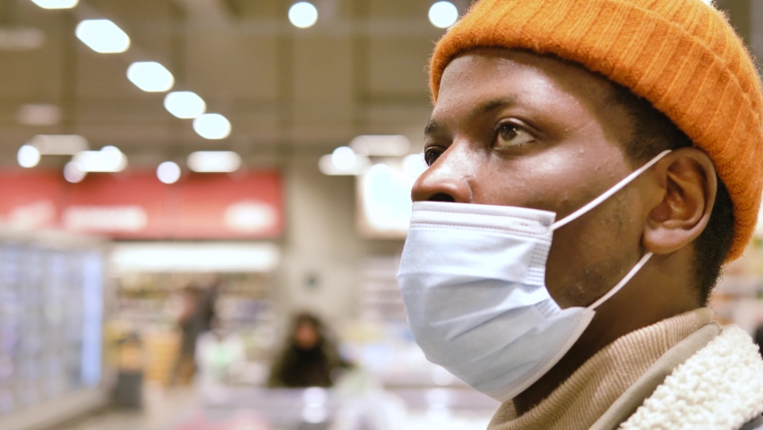 Black man in hat and face mask walks along grocery store and looks around choosing products under electric light close view