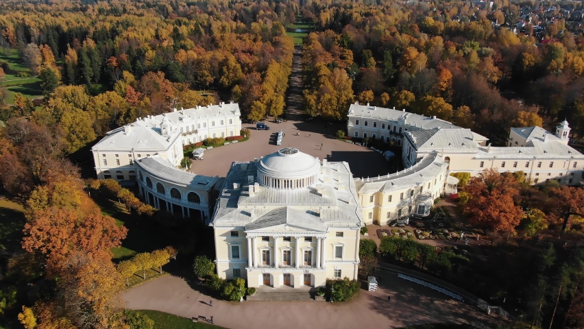 Semicircular white building of Pavlovsk palace among large city park with terracotta leaves on trees in autumn aerial panorama