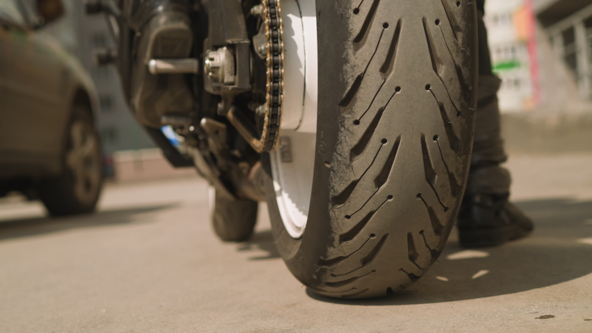 Driver in black moto suit and boots rides modern motorcycle along parking site against blurry dwelling building on sunny day backside view slow motion