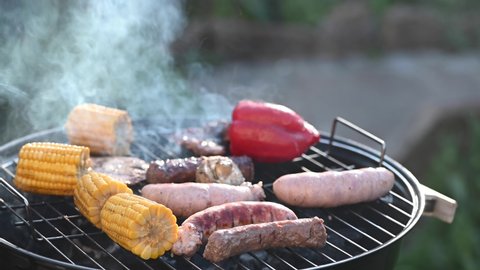 Fried sausages with corn and red pepper on a round black grill, covered with a lid, with smoke with a landscape in the country