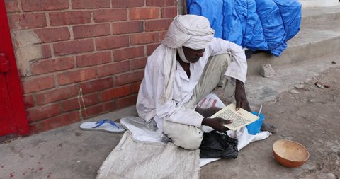NIMA, GHANA - 23 OCT 2021: Muslim man on road reading Torah Accra Ghana. Muslim man begs on the street while reading scripture and  Koran Torah. Morning prayer Accra Ghana. Islam religion.