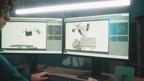 Man in glasses using computer to create 3D model of futuristic aircraft for videogame while working in home office at night