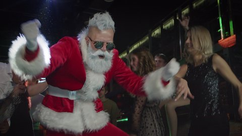 Modern energetic Santa Claus in red suit and cool sunglasses dancing and waving hands in center of dancefloor among flashes of light and carefree smiling men and women, celebrating Christmas in club.