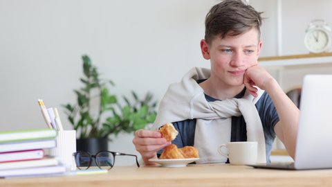 Home education teenage kids. Positive teen boy bites piece of croissant having break and listening to teacher via laptop at table in room closeup 4k video