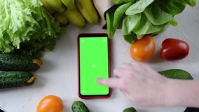 Smartphone with green screen surrounded by farm fruits and vegetables on white table. Unrecognizable person is scrolling through app. Online shopping for grocery products and ordering food delivery - Powered by Shutterstock - Get 15% off with code: PIKWIZARD15