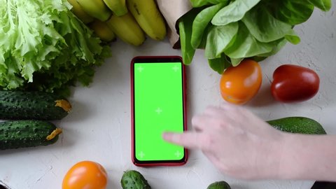 Smartphone with green screen surrounded by farm fruits and vegetables on white table. Unrecognizable person is scrolling through app. Online shopping for grocery products and ordering food delivery