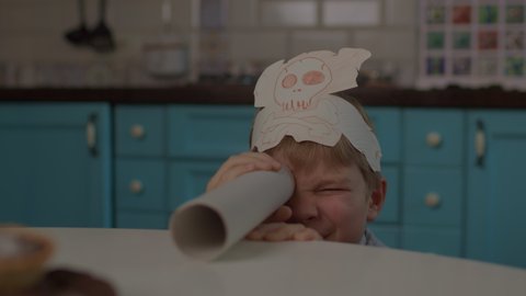 Kid playing pirate's game with paper telescope and headwear with skull and crossbones. Preschool boy searching for sweets with pirate's telescope.