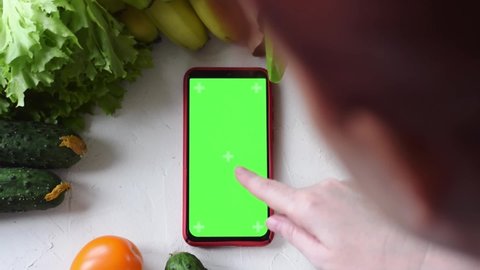 Smartphone with green screen surrounded by farm fruits and vegetables on white table. Unrecognizable person is scrolling through app. Online shopping for grocery products and ordering food delivery
