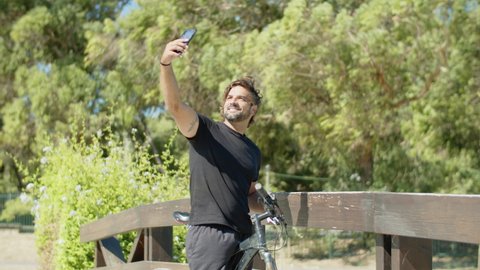 Vertical motion of biker taking selfie on phone in summer park. Medium shot of happy man standing on bridge with bicycle, looking into mobile camera, showing shaka sign. Photoshoot, sport concept