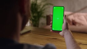 Handheld Camera: Young Man at Home Holding Chroma Key Green Screen Smartphone Watching Content Without Touching or Swiping. Browsing Internet, Watching Content, Videos