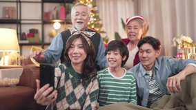 Smiling asian big family with kid on christmas party sit on couch in living room make self-portrait video on cell together, happy parents with small children have fun videocall on smartphone at home 