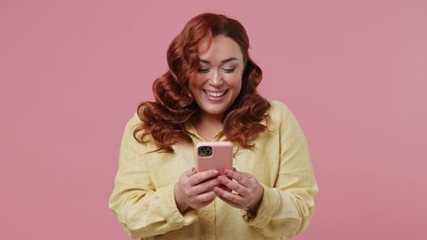 Happy charming vivid bright young ginger chubby overweight woman 20s wears yellow shirt hold using mobile cell phone typing browsing chatting send sms isolated on plain pink background studio portrait