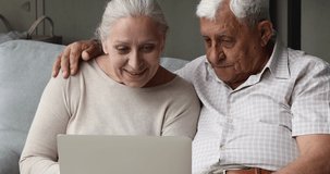 Middle aged couple retirees study basics of using modern tech laptop learn to work in computer app together on couch at living room. Hugging older grandparents discuss online purchase before pc screen
