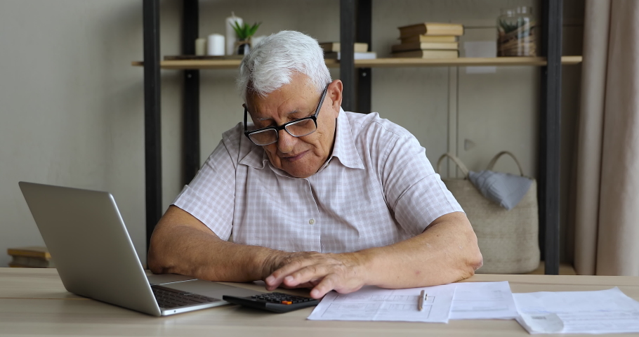 Retired man study financial papers at home office check information on calculator before pay online using laptop. Elderly male on pension do home banking work review mortgage loan insurance payments