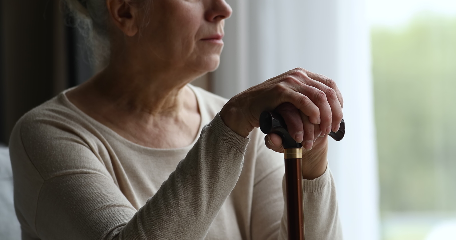 Cropped close up of depressed hoary older adult female living with physical disability sit by window alone hold stick. Retired old woman with walking difficulties look at distance feel weak hopeless | Shutterstock HD Video #1082309506