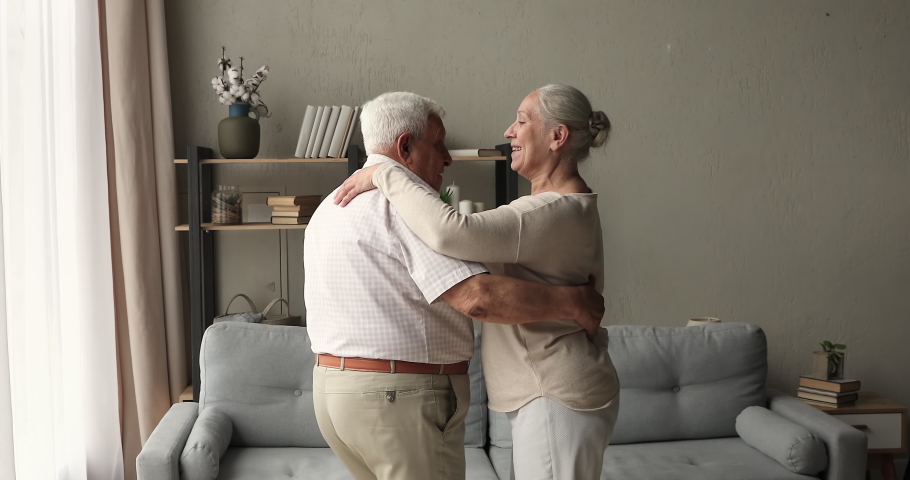 Happy carefree older adult married couple enjoy romantic dance together at living room of own flat apartment celebrate anniversary. Energetic healthy aged spouses waltzing remembering days of youth Royalty-Free Stock Footage #1082309560