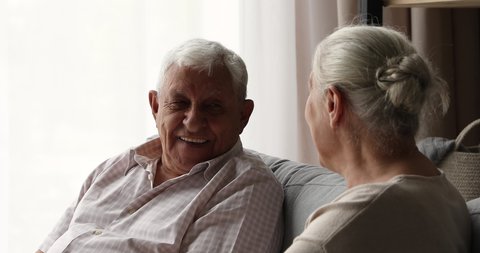 Positive older age male female on pension spouses neighbors friends chat on comfy couch engaged in pleasant dialogue relax at home together. Elderly couple talk discuss interesting topics having fun