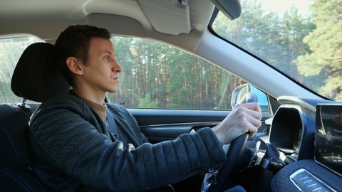 Brunet driver wearing dark sheepskin coat holds steering wheel confidently and drives foreign car along forest road on sunny autumn day closeup
