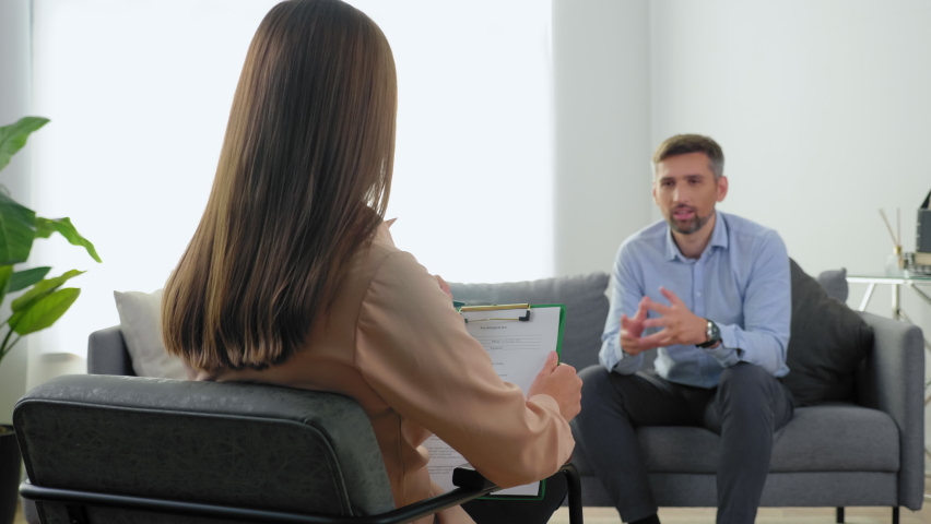 Woman psychologist taking notes on clipboard and listens client at psychotherapy session. Unfocused adult man patient with mental health problems after covid-19 sitting on couch talking to therapist | Shutterstock HD Video #1082310793