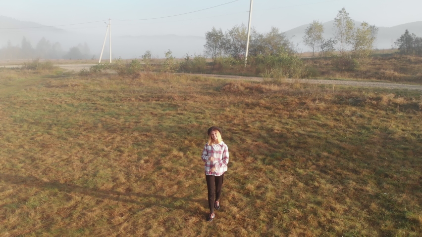 Aerial drone view of young woman walk on grass in nature in meadow near rural village with amazing mountains and cloudy background. People travel and enjoy nature, countryside fields, valley, forest