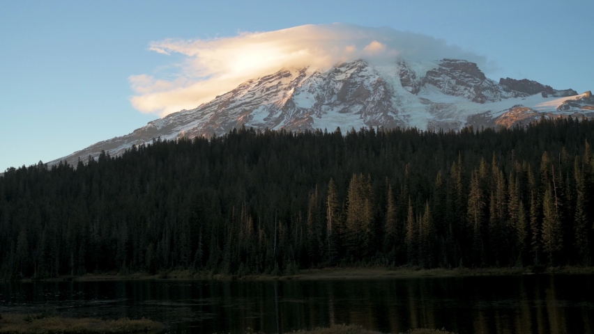 4K video of the PNW volcano Mount Rainier with its ice and snow ridges rising above green forest and a lake in Washington State at sunset