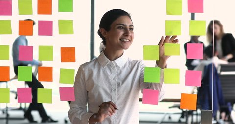 Young Indian woman write down step by step instructions for different tasks, strategizing new projects using sticky notes placed on transparent wall. Subdivide goals, make important reminders concept
