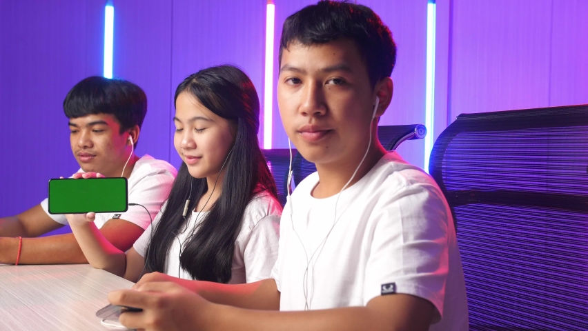 Team Of Asian Gamers Sitting On Chair And Pointing To Mock Up Green Screen Mobile Phone
