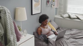 Medium slowmo shot of cheerful young African-American woman in homewear spending weekend morning at home, video chatting with friend on laptop sitting on soft bed at cozy apartment