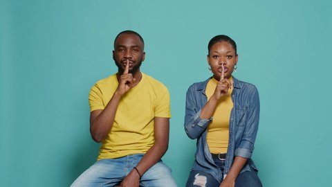 Man and woman doing silence gesture with index finger on mouth, asking for secrecy and quiet. Couple showing sign of privacy to keep secret and request to shut up, sitting over blue background
