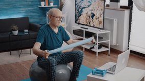 Pensioner stretching resistance band to work arms muscles while watching video of training lesson on laptop. Elder man using elastic belt and computer, sitting on fitness toning ball.